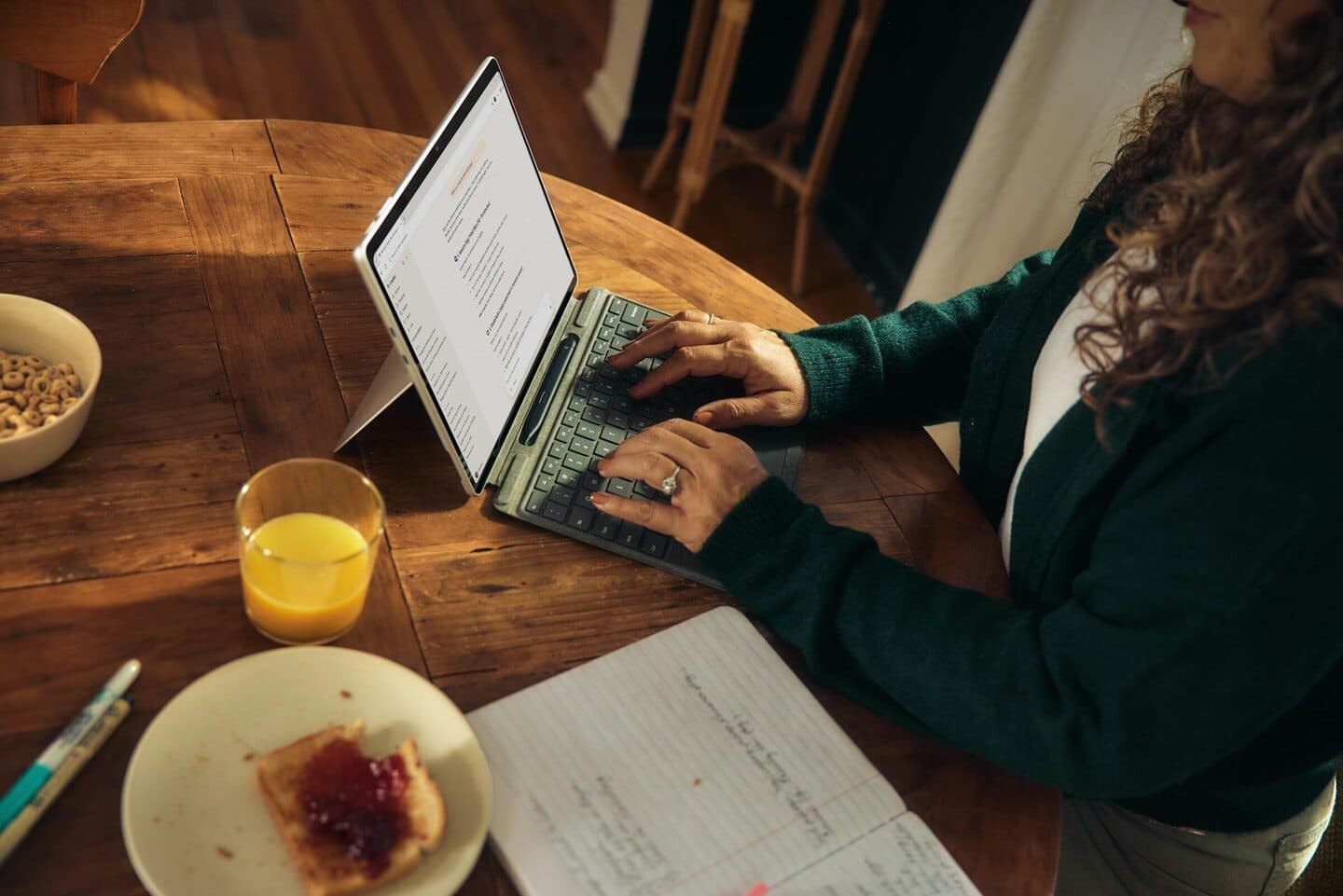 Frau tippt an einem Holztisch auf einem Tablet mit Tastatur, daneben Frühstück und Notizen