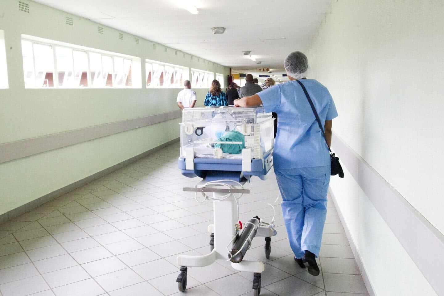 Pflegekraft in blauer Kleidung schiebt einen Inkubator durch einen Krankenhausflur
